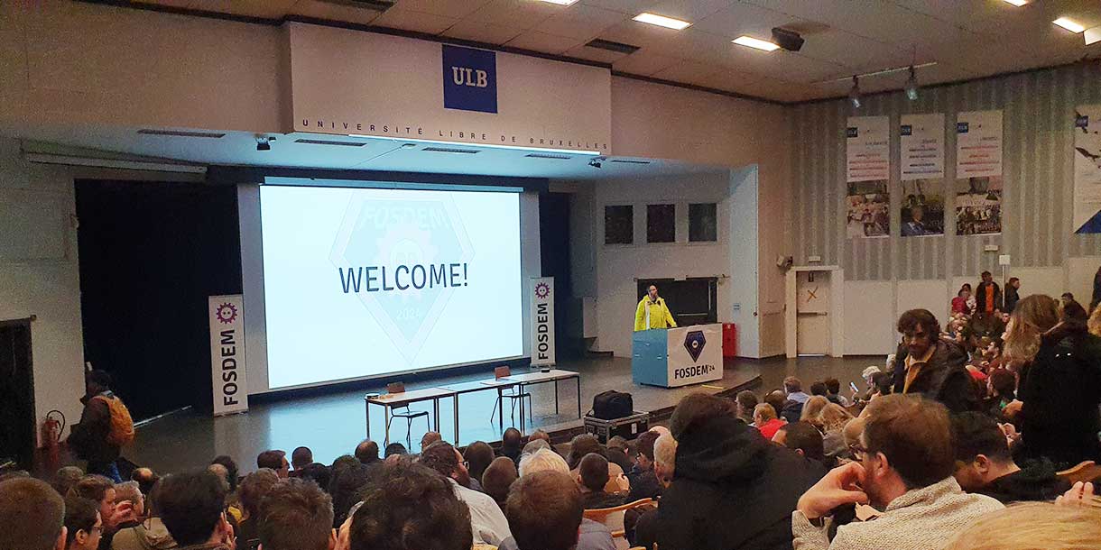 FOSDEM 2024, konferensen för fri och öppen källkod vid Université Libre de Bruxelles, med "Welcome" på storbildsskärmen och en fullsatt aula.